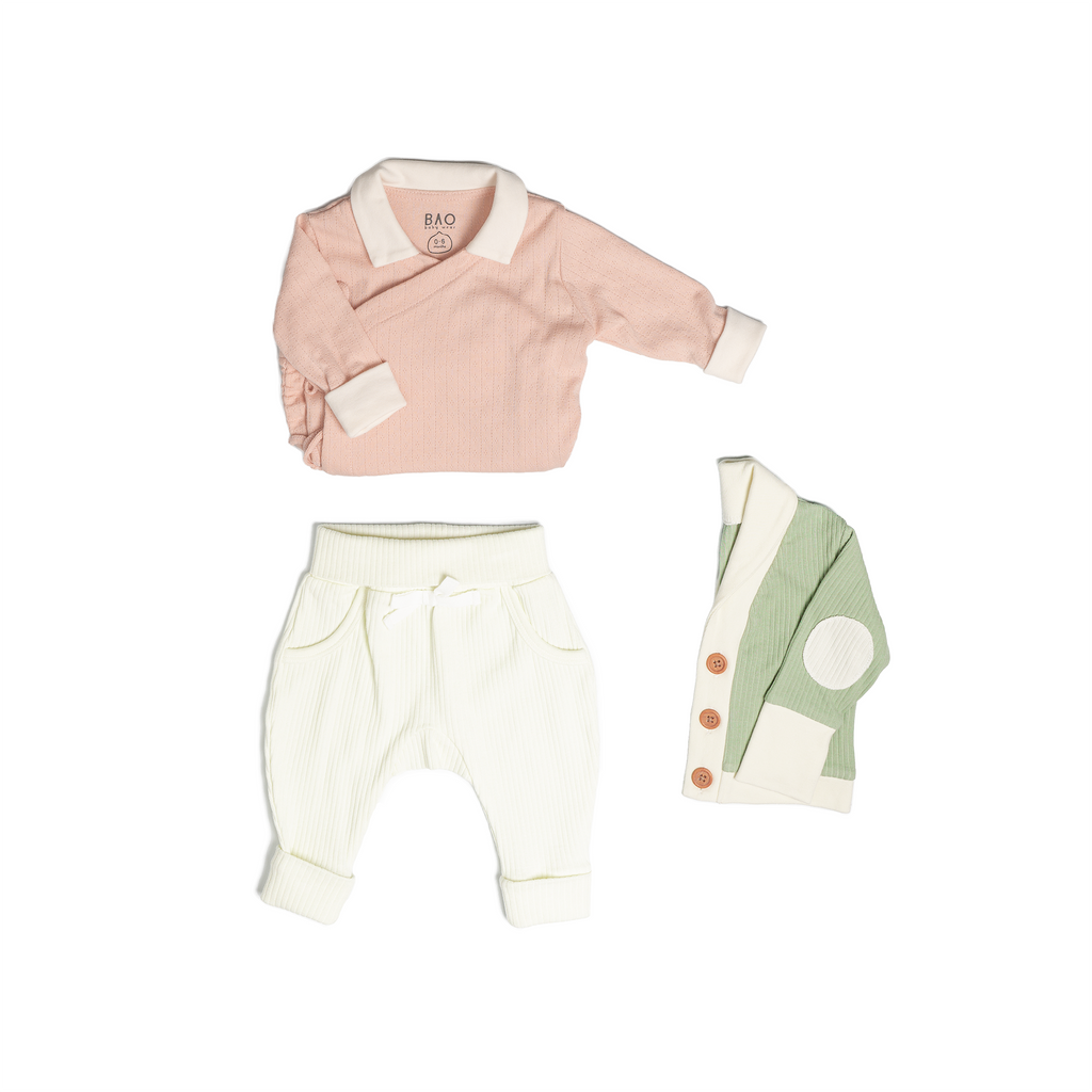 The newborn essentials bundle, consisting of a kimono bodysuit in a peach pointelle patterned cotton, paired with lounge pants in vanilla and ribbed cotton and a cardigan in matcha ribbed cotton.
