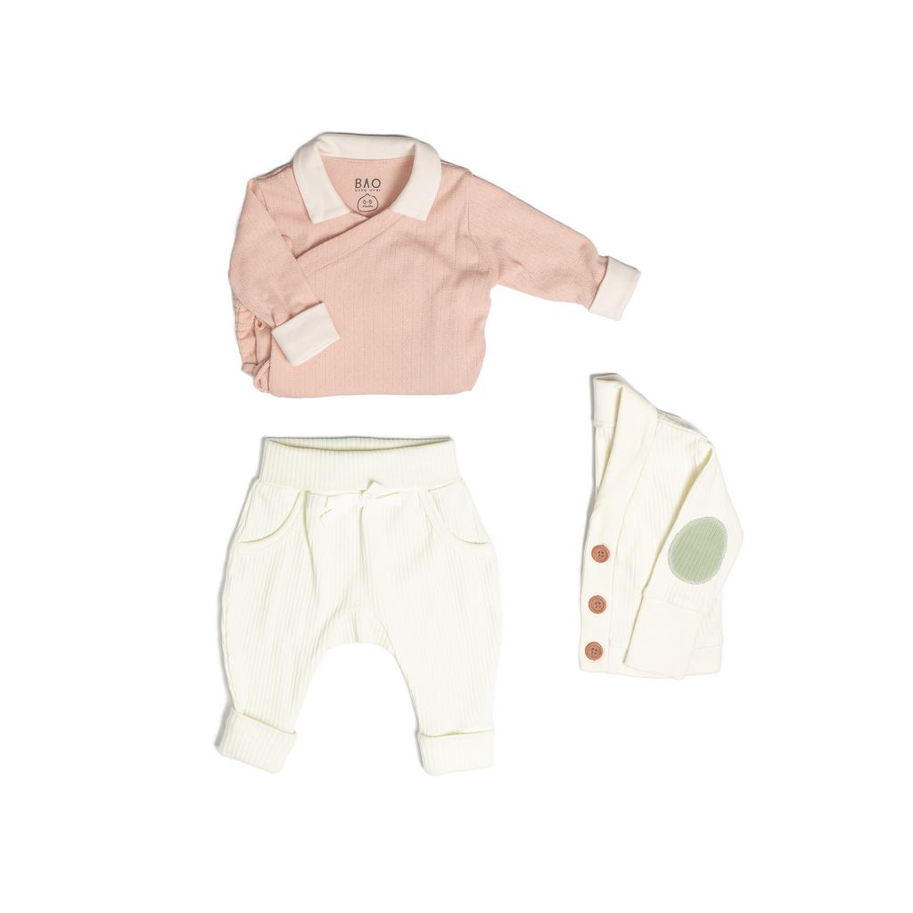 The newborn essentials bundle, consisting of a kimono bodysuit in a peach pointelle patterned cotton, paired with lounge pants in vanilla and ribbed cotton and a cardigan in vanilla ribbed cotton.